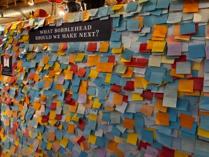 A wall of post-it notes from visitors answering the prompt "what bobblehead should we make next?"